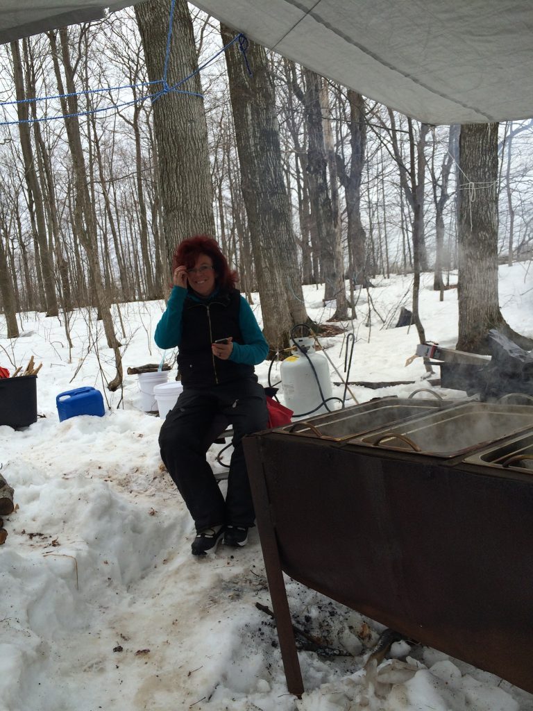woman reading her phone in front of an evaporator which is used for maple syrup production. She is under a tarp in the woods